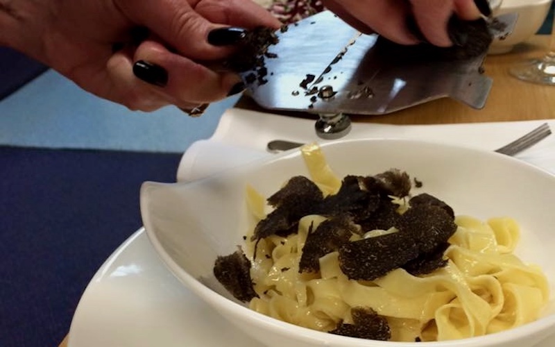 Hand using stainless steel truffle grater to grate a black truffle over a white bowl of fettucine with black truffle